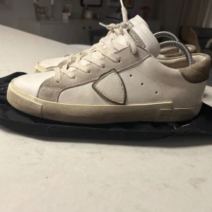 Philippe Model vita sneakers i skinn - Snygga vita sneakers från Philippe Model med mörk gröna mockadetaljer och klassisk snörning. Skorna har en rund tå, platt sula och ikonisk sköld på sidan. Tillverkade i skinn och mocka för en lyxig känsla. Perfekta för dig som gillar stilrena och trendiga sneakers. Produkten är sparsamt andvänd och i mycket bra skick.