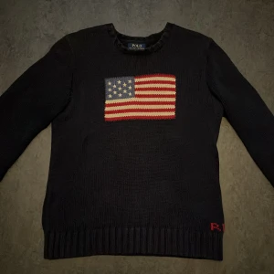 Mörkblå stickad tröja Polo Ralph Lauren - Snygg mörkblå stickad tröja från Polo Ralph Lauren med amerikansk flagga framtill och RL-broderi vid nederkanten. Klassisk rund halsringning och långa ärmar. Perfekt för dig som gillar preppy stil och vill sticka ut lite extra. Storlek S/M