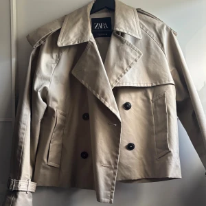 Beige kort trenchcoat från Zara XS - GÅR EJ NER I PRIS Kort trenchcoat från Zara i beige med dubbelknäppning, stora slag och bälte vid ärmsluten. Jackan har en loose passform och är tillverkad i bomullsmix. Perfekt för vår och höst när du vill ha en clean och stilren look.