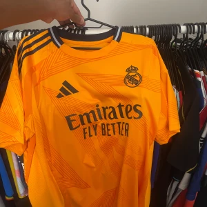 Real Madrid 2024 Tredjeställ - Snygg orange Real Madrid fotbollströja från Adidas med Rodrygo och nummer 11 på ryggen. Tröjan har svarta detaljer, klubbmärke och sponsortryck framtill samt klassiska Adidas-ränder på axlarna. Tillverkad i lätt och ventilerande material.