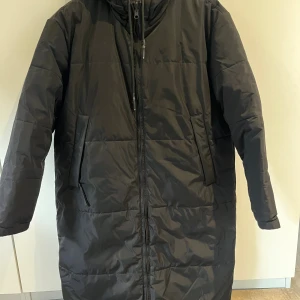 Svart lång pufferjacka från Brixtol - Lång svart pufferjacka från Brixtol med huva och dragkedja framtill. Jackan har två stora fickor på sidorna och quiltad design. Perfekt för kalla dagar och har en clean, minimalistisk look. Du kan vända den så den blir både svart och mörkblå.