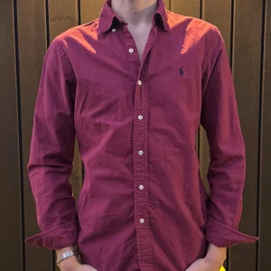 Vinröd skjorta från Ralph Lauren - Säljer en vinröd skjorta från Ralph Lauren med klassisk krage och knappar framtill. Skjortan har lång ärm och den ikoniska broderade loggan på bröstet. Tillverkad i bomull och passar perfekt för dig som vill ha en stilren look.