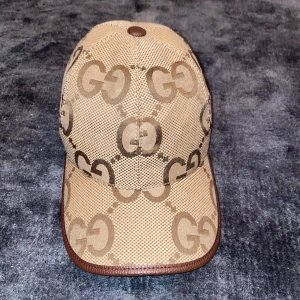 Beige keps från Gucci med GG-mönster - Snygg beige keps från Gucci med klassiskt GG-monogram i brunt över hela kepsen. Kepsen har bruna skinn-detaljer och justerbart spänne bak. Tillverkad i canvas med läderdetaljer för en lyxig känsla. Perfekt för dig som vill sticka ut med en ikonisk accessoar. Denna är den bästa a kopian man kan hitta på tanke på kvaliten.