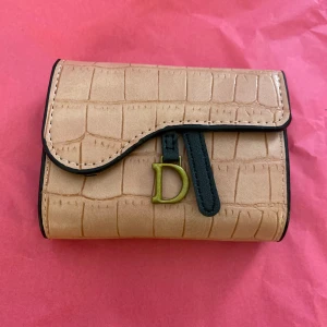 Rosa plånbok med krokoprägling och D-detalj - Snygg rosa plånbok med krokodilpräglat mönster och guldfärgad D-detalj framtill. Plånboken har svart kant, tryckknappsstängning och flera fack för kort och sedlar. Perfekt accessoar för dig som gillar stilrena detaljer och lyxig känsla.