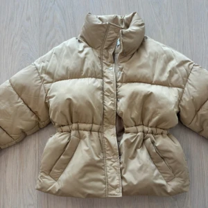 Beige pufferjacka från NA-KD - Snygg beige pufferjacka från NA-KD med hög krage, dragkedja och tryckknappar framtill. Jackan har markerad midja med resår och två stora fickor. Perfekt för kalla dagar och ger en cool, trendig look.