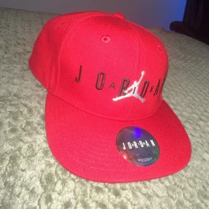 Röd Jordan keps med broderad logga - Snygg röd keps från Jordan med svart broderad text och den klassiska Jumpman-loggan i vitt framtill. Kepsen har platt skärm och justerbar passform. Tillverkad i bomull och perfekt för dig som vill ha en sportig och trendig look.