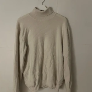 Beige polotröja i mjuk ull - Snygg beige polotröja med ribbad krage och muddar. Tröjan är långärmad och gjord i mjuk ull, perfekt för kyliga dagar. Enkel och clean design som är lätt att matcha med andra plagg. Passar dig som gillar stilrena och bekväma tröjor.