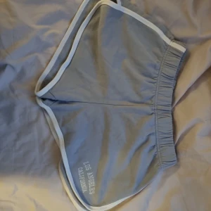Ljusblå shorts från Brandy Melville - Säljer ett par ljusblå shorts från Brandy Melville med vit kant och resår i midjan. På ena benet står det 'Los Angeles California' i vitt. Perfekta för sommaren och riktigt sköna att ha på sig.