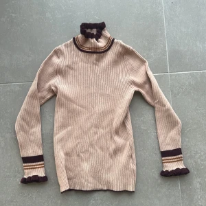 Rosa ribbad polotröja  - Snygg rosa ribbad polotröja med mörkbruna och ljusbruna detaljer vid hals och ärmslut. Polokragen och ärmsluten har volangkant som ger en extra touch. Perfekt för lager-på-lager och passar till både jeans och kjol.