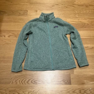 Grå fleecejacka från Patagonia L - Mysig grå fleecejacka från Patagonia i storlek L. Jackan har hel dragkedja framtill, hög krage och två sidofickor med dragkedja. Tillverkad av 100% återvunnen polyester, perfekt för kyliga dagar.