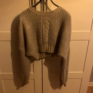 Beige croppad stickad tröja NA-KD - Supermysig croppad stickad tröja från NA-KD i beige. Tröjan har lång ärm, bred ribbad mudd och ett snyggt flätmönster framtill. Perfekt för lager-på-lager och chill höst- eller vinterstil.