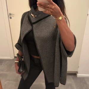 Trekvartsärm poncho/ cape  - Trekvartsärm poncho/cape med snyggt guldspänne vid halsen. Jackan har ett stickat mönster och svarta detaljer längs kanterna 😍