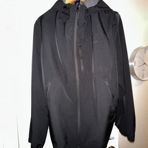 Svart vindjacka från GANT, strl M - Svart vindjacka från GANT med huva och flera dragkedjefickor. Jackan har justerbar snörning vid halsen och är tillverkad i slitstark polyester. Perfekt för dig som gillar stilren och funktionell design. Passformen är normal och jackan har långa ärmar.