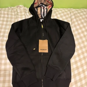 Svart Burberry hoodie med dragkedja - Svart hoodie från Burberry med klassiskt rutigt foder i huvan och guldfärgade detaljer på snörningen. Tröjan har dragkedja framtill, ribbade muddar och stor huva. Perfekt för dig som gillar stilrena och lyxiga streetwear-plagg.