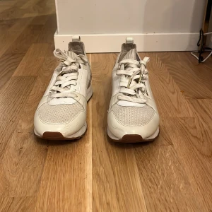 Vita sneakers från Michael Kors - Snygga vita sneakers från Michael Kors med guldiga detaljer och MK-logga baktill. Skorna har snörning, perforerade partier och stickade inslag för en sportig look. Perfekta för dig som gillar stilrena och trendiga sneakers med exklusiv känsla. Finns mycket slitning på skorna därför säljs det billigt 