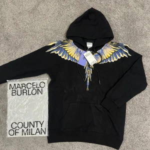 Marcelo Burlon svart hoodie med vingar - Svart hoodie från Marcelo Burlon County of Milan med ikoniskt vingtryck i blått och gult över axlar och rygg. Tröjan har huva med snörning och känguruficka framtill. Perfekt statement-plagg för dig som vill sticka ut.