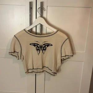 Beige croppad topp med fjäril Divided - Trendig croppad topp från H&M Divided i beige med svart tribal-fjäril på bröstet. T-shirten har kontrastsömmar i svart och korta ärmar. Perfekt för dig som gillar en edgy och cool stil.