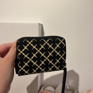 Svart och beige plånbok från By Malene Birger - Snygg plånbok från By Malene Birger i svart och beige med ikoniskt grafiskt mönster. Plånboken har dragkedja runtom och flera fack för kort och sedlar. Tillverkad i tåligt skinnliknande material och perfekt storlek för att ha med sig överallt.