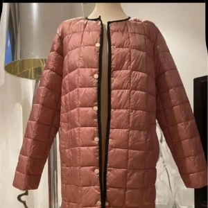 Rosa quiltad pufferjacka med knappar - Lång, quiltad pufferjacka i matt rosa med svarta detaljer längs kanterna. Jackan har rund halsringning, tryckknappar framtill och raka ärmar. Den är lätt vadderad och har en stilren, minimalistisk look som passar perfekt till kyliga dagar.