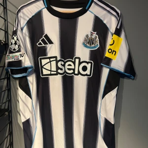 Newcastle United Isak 14 Adidas tröja - Snygg Newcastle United matchtröja från Adidas med Isak 14 på ryggen. Tröjan har klassiska svarta och vita ränder, blå detaljer och klubbmärke på bröstet. Kortärmad modell i lätt polyester, perfekt för fotboll eller häng med polarna. Helt ny tröja med tagg. En replika