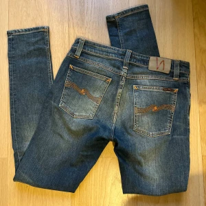 Nudie Jeans - Säljer ett par blå Nudie jeans med klassiska orange sömmar och vågigt broderi på bakfickorna. Jeansen har normal midja, och är tillverkade i mjukt denimtyg. Grymt skick och inga som helst defekter!