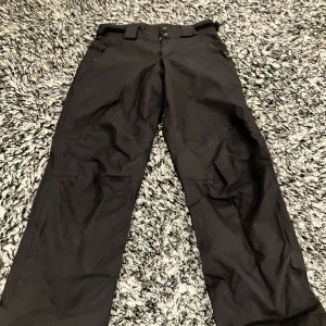 Svarta täckbyxor från Neomondo - Svarta täckbyxor från Neomondo med justerbar midja med kardborre och bälteshällor. Byxorna har dragkedja i sidan och dubbla knappar framtill. Perfekta för kalla dagar och utomhusaktiviteter. 