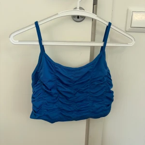 Blå topp - Säljer en blå topp med smala axelband och snygga rynkade detaljer framtill. Toppen har en croppad passform och är perfekt för varma dagar eller som layering-piece. Materialet känns mjukt och stretchigt.