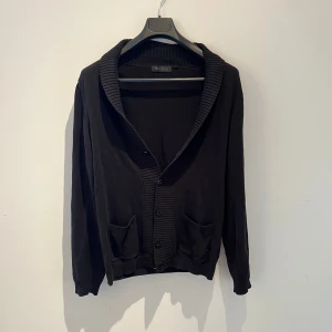 Svart kofta från Riley med fickor - Svart kofta från Riley med ribbad sjalkrage och knäppning framtill. Tillverkad i mjukt material som passar perfekt för lager-på-lager. Långärmad och stilren design som funkar till många outfits. Inga defekter, bara hör av er för fler bilder eller andra frågor