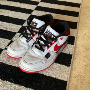 Nike Air Force 1 Low vit/röd - Säljer ett par Nike Air Force 1 Low med vit ovandel i skinn, röda detaljer och svart foder. Skorna har svart snörning, perforerad tå och klassisk röd swoosh på sidorna. Sulan är vit, röd och grå med Nike-logga undertill. Perfekta för dig som gillar streetstyle.