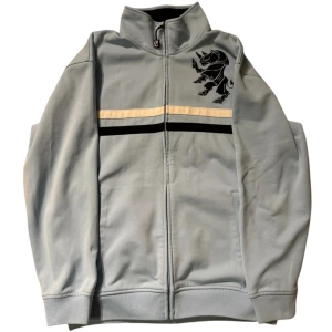 Ljusblå zipup från Ecko Unltd. - Säljer en ljusblå zipup från Ecko Unltd. med dragkedja och ett coolt tryck av en noshörning på bröstet. Jackan har svarta och vita ränder över bröstet och en hög krage. Perfekt för en avslappnad stil. Lite typ noppror på ena ärmen men går enkelt bort annars märks det knappt