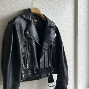 Svart skinnjacka från Zara - Snygg svart skinnjacka från Zara med klassisk bikerstil, silvriga dragkedjor, axelklaffar och bälte i midjan. Jackan har flera fickor med dragkedja och coola detaljer som ger en edgy vibe. Perfekt för dig som vill ha en tidlös och trendig look. Något oversized