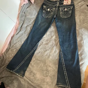 True Religion bootcut jeans med kontrastsömmar - Säljer ett par mörkblå bootcut jeans från True Religion med tydliga vita kontrastsömmar och ikoniska fickdetaljer bak. Jeansen har låg midja och klassisk femficksdesign. Materialet är jeans i bomull och de har en snygg tvättad look. Dom är knappt använda och passar på mig som är 1,67 och har till vanligt S i jeans 
