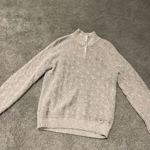 Grå stickad tröja med half zip - Säljer en grå stickad tröja med snyggt kabelmönster och half zip vid halsen. Tröjan har långärmade och ribbade muddar vid ärmslut och nederkant. Perfekt för dig som gillar klassisk stil med en modern twist.