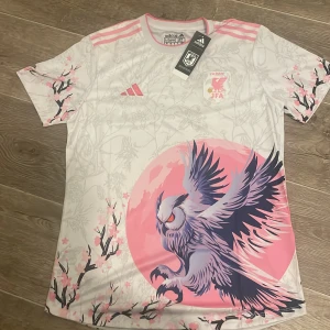 Japan JFA fotbollströja Adidas M/S - PASSAR SOM SJapan JFA fotbollströja från Adidas i vitt och rosa med snyggt körsbärsblom-mönster och stor fågelgrafik på framsidan. Tröjan har korta ärmar, rosa detaljer och är tillverkad i lätt, ventilerande material. Perfekt för dig som vill sticka ut på planen. 