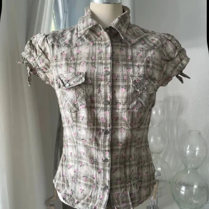 Kortärmad rutig skjorta med blommor - Snygg kortärmad skjorta med beige och grå rutigt mönster, dekorerad med små rosa blommor. Skjortan har knappar framtill, två bröstfickor med lock och knytband på ärmarna. Materialet känns lätt och luftigt, perfekt för varmare dagar. Figurnära passform och klassisk krage.