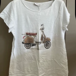 Vit t-shirt med vespa-tryck Marta du Château - Vit t-shirt från Marta du Château i 100% bomull. Framsidan har ett coolt tryck av en vespa med små nitar som detalj. T-shirten har rund hals och korta ärmar, perfekt för en chill och avslappnad stil. Lite mindre i storlek 