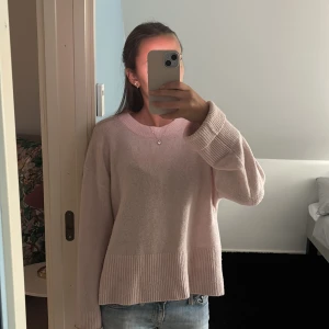 Ljusrosa stickad tröja med ribbade detaljer - Mysig ljusrosa stickad tröja med breda ribbade muddar vid ärmslut och nederkant. Tröjan har en rund halsringning och en loose passform som ger en chill vibe. Perfekt att matcha med jeans för en enkel och snygg look.