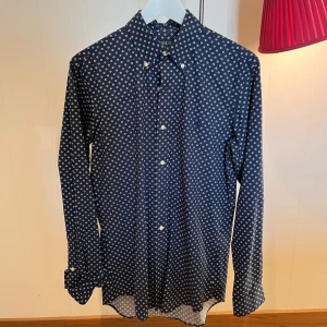 Mörkblå mönstrad skjorta Polo Ralph Lauren - Snygg mörkblå skjorta från Polo Ralph Lauren med custom fit. Skjortan har ett vitt blommigt mönster över hela tyget, klassisk krage och vita knappar framtill. Långärmad och tillverkad i mjuk bomull som känns skön mot huden. Perfekt för dig som gillar stilrena detaljer.