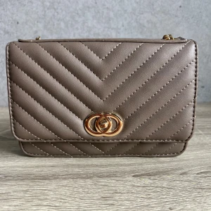Beige axelväska med guldkedja - Snygg beige axelväska i quiltad design med guldfärgad kedja och dekorativt spänne framtill. Väskan har flera fack, inklusive ett med dragkedja. Perfekt storlek för det viktigaste.