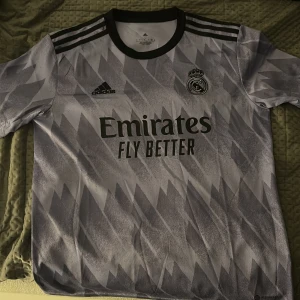 Real Madrid Adidas fotbollströja L - Snygg Real Madrid fotbollströja från Adidas i storlek L. Tröjan är kortärmad med svart krage och axelränder, har ett grått och vitt geometriskt mönster samt broderade loggor för både Adidas och Real Madrid. Tillverkad i lätt och ventilerande polyester.