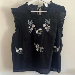 Svart blommig blus  - Svart blus från H&M Divided i storlek 38. Blusen har broderade blommor i vitt och ljusgult framtill, volangdetaljer vid axlarna och är ärmlös. Rund hals med knytning bak i nacken.