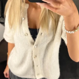 Vit stickad kofta med pärlknappar - Superfin vit stickad skjorta med korta ärmar och snygga pärlknappar framtill. Koftan har en mjuk och fluffig känsla, perfekt för dig som gillar en chill och trendig look. Passar dig som vill ha något enkelt men ändå stilrent.💕