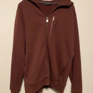 Vin röd hoodie från Sail Racing L - Säljer en vin röd hoodie från Sail Racing i storlek L. Tröjan har dragkedja framtill, en extra bröstficka med dragkedja och en huva med knappdetalj bak. Mjuk insida och ribbade muddar vid ärmslut och nederkant. Perfekt för chill dagar.