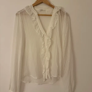 Vit blus med volang från Stradivarius - Snygg vit blus från Stradivarius i storlek S. Blusen har tunna vertikala ränder, v-ringning med volangdetaljer och knappar framtill. Långärmad och tillverkad i ett lätt, transparent tyg som ger en fräsch och luftig känsla.