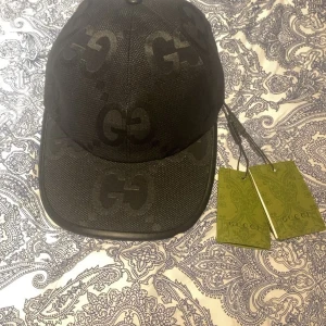 Gucci Keps - Svart keps från Gucci med klassisk böjd skärm och tydliga GG-loggor ton-i-ton över hela kepsen. Tillverkad i ett slitstarkt polyestertyg och justerbar baktill. Märkets etikett och gröna originaltags medföljer. Snygg accessoar för dig som gillar exklusiva streetwear-vibbar. Den har en prislapp inte 2