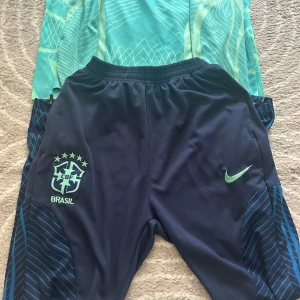 Nike Brasil träningsbyxor blå - Säljer ett par mörkblå Nike Brasil träningsbyxor med elastisk midja och grönt CBF Brasil-emblem på vänster ben. Snygga detaljer med blå ränder längs sidorna och en grön Nike-logga på höger ben. Perfekta för fotboll eller träning. Skriv privat om ni vill ha tröjan med så kan man diskutera pris.