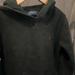 Svart hoodie från Polo Ralph Lauren - Svart hoodie från Polo Ralph Lauren med klassisk röd broderad logga på bröstet. Tröjan har huva, känguruficka och är tillverkad i mjuk bomull. Perfekt för en chill och stilren look.