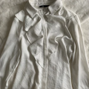 Vit blus med volang från Zara - Supersnygg vit blus från Zara med volangdetaljer framtill och pärlknappar vid hals och ärmslut. Klassisk krage och långärmad modell i ett mjukt, lätt material som känns lyxigt. Perfekt för dig som gillar stilrena men ändå unika plagg.