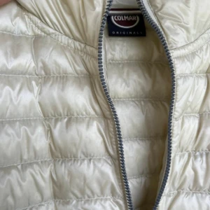 Beige dunjacka från Colmar - Snygg beige dunjacka  från Colmar Originals med dragkedja framtill och två dragkedjeförsedda fickor. Västen har en stilren design med lätt glansigt material och Colmar-logga på ärmen. Perfekt för lager-på-lager och streetstyle.  ITALIENSK 42a