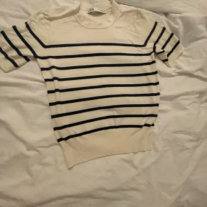 Randig kortärmad tröja från Zara - Säljer en vit och marinblå randig tröja från Zara i storlek S. Tröjan har korta ärmar, rund halsringning och är tillverkad i ett mjukt, stickat material. Perfekt för en chill och stilren look.